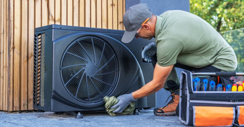 A technician wearing blue gloves, a grey hat, and a green shirt is wiping down a black HVAC system outside.