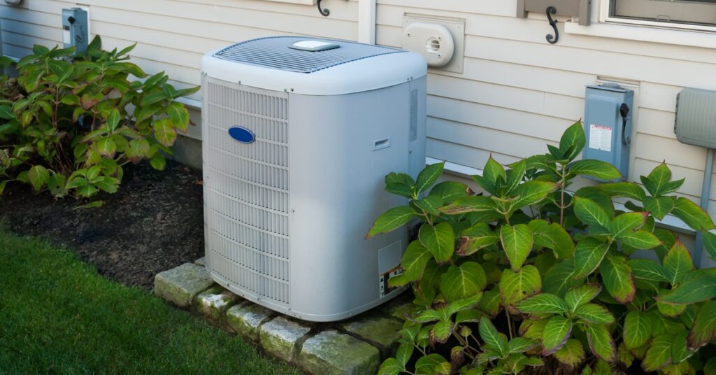 An outdoor heating and air conditioning inverter unit mounted on a home’s exterior wall beside a window.
