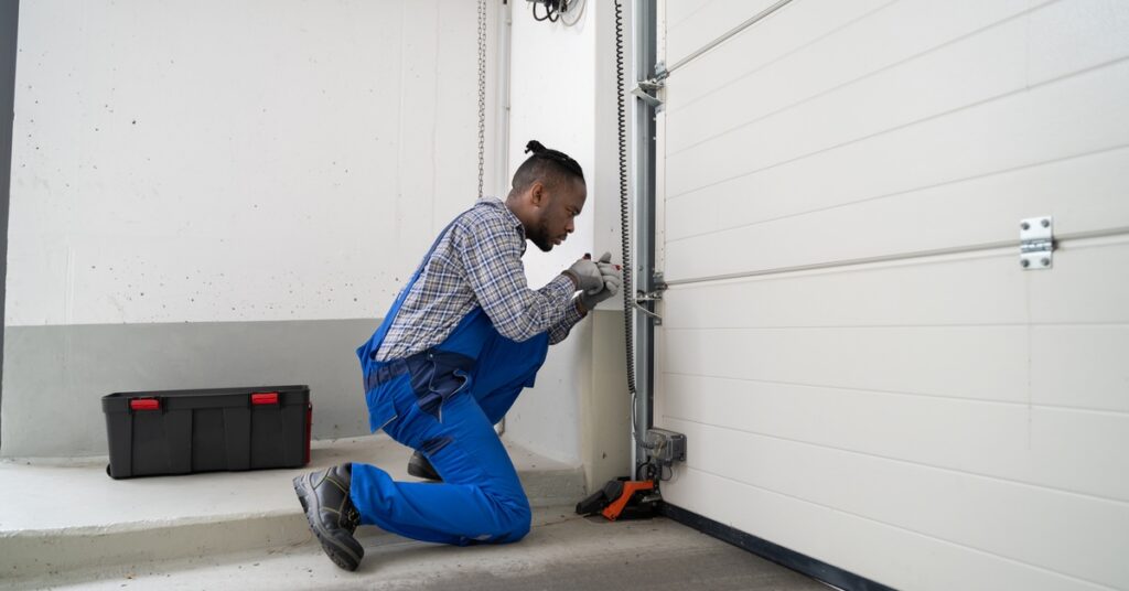 a man kneeling beside a garage door track while using tools to work on a door system inside a residential garage.