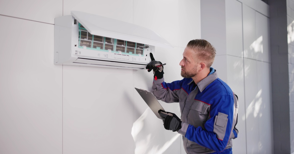 A man in a blue and gray work uniform holding a clipboard inspects an open wall-mounted air conditioner.