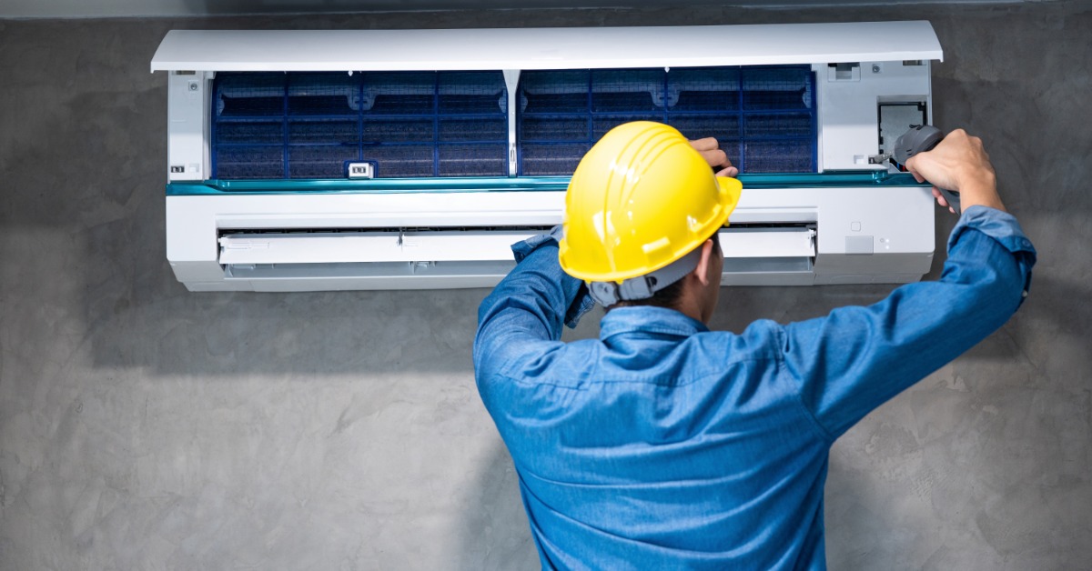 A man wearing a yellow hard hat uses a screwdriver on an open wall-mounted air conditioner against a gray wall.