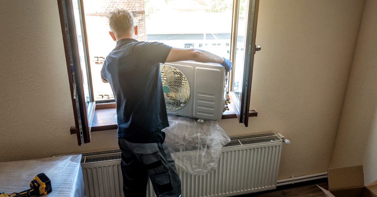 A man standing by an open window carries an outdoor air conditioner unit into a room with tools and boxes.