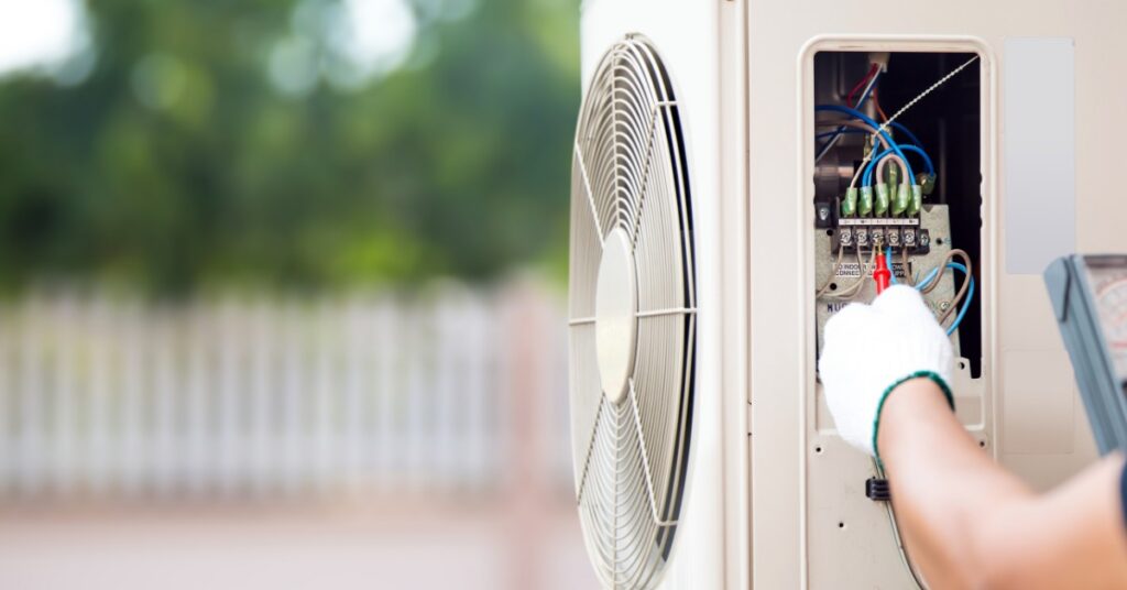 A gloved hand using a meter tests an open outdoor air conditioner panel with red and black probe leads.