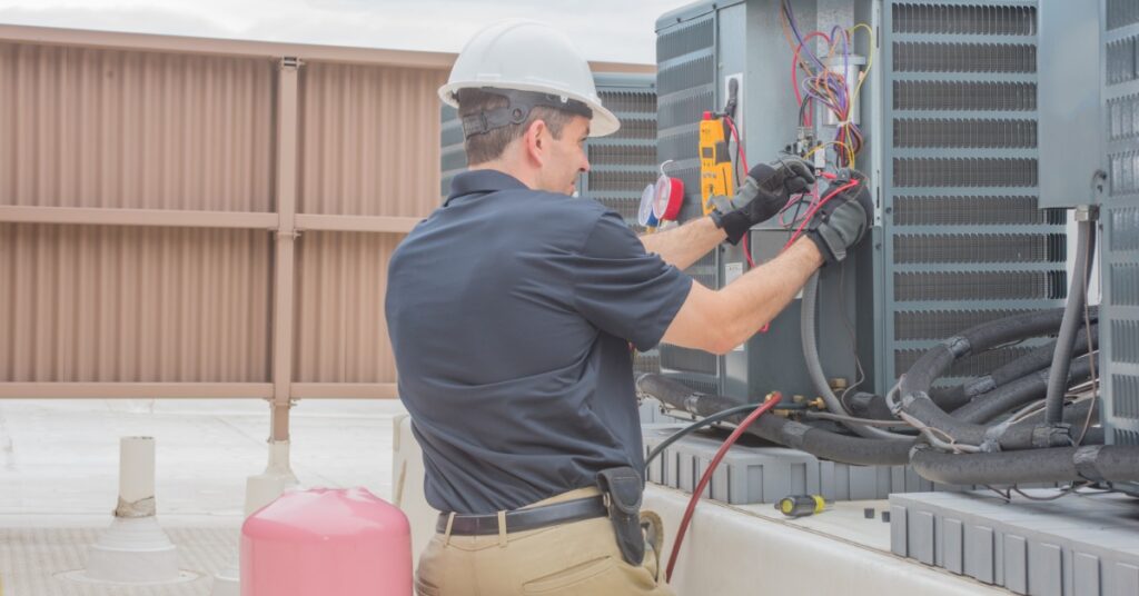 A person in a hard hat checks an open rooftop condenser unit with test leads, tools, and a cloudy blue sky.