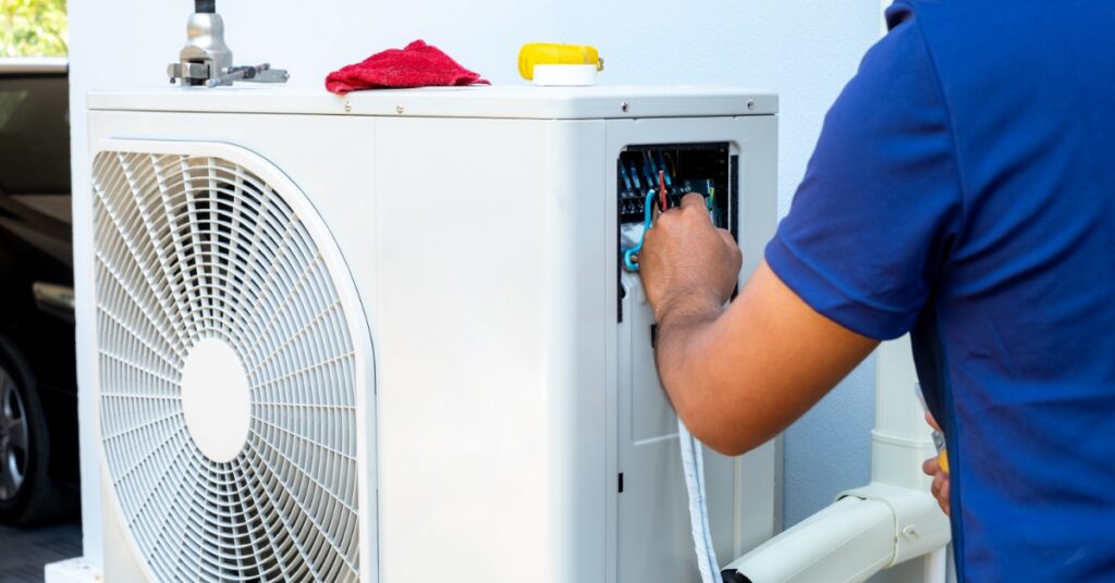 A person in a red uniform works from a ladder on a wall-mounted air conditioner unit under a blue sky.