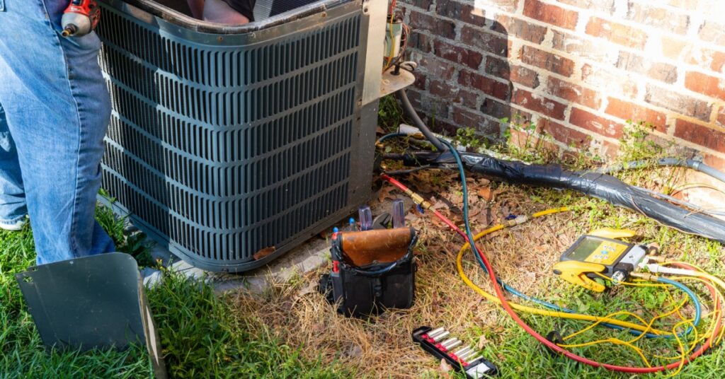 A partially visible person working inside an outdoor air conditioning unit beside hoses and tools near a brick wall.