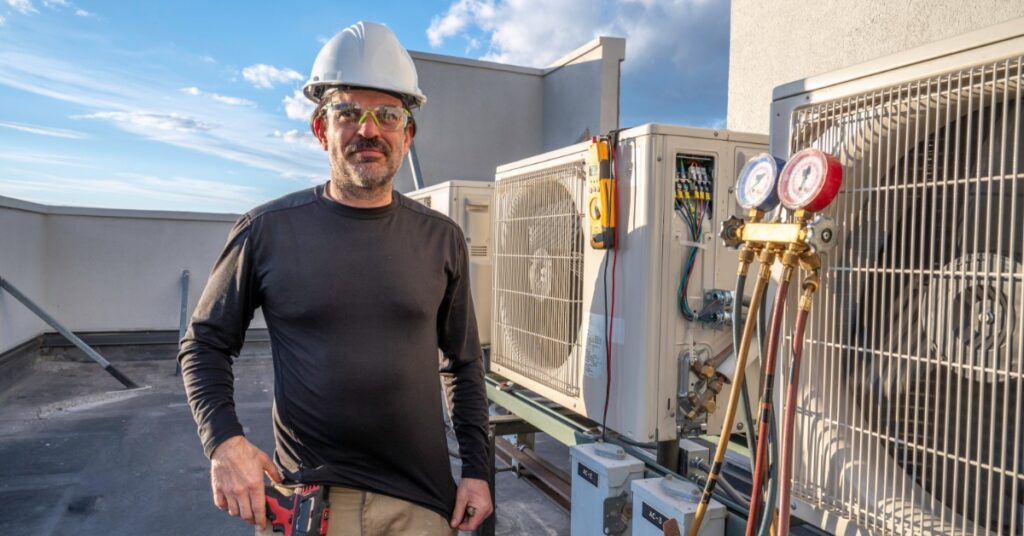 A man wearing a white hard hat and safety glasses standing on a rooftop beside outdoor air conditioning units.