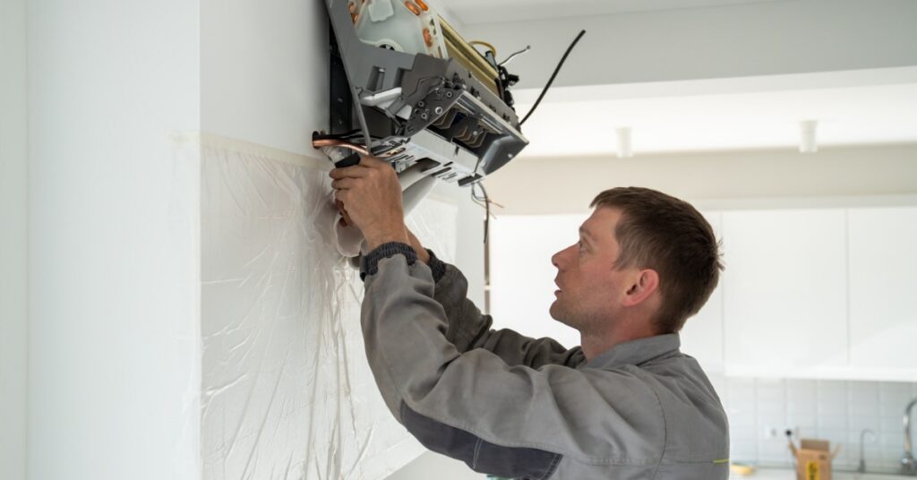 A man in a gray work shirt working on an open wall-mounted air conditioning unit in a bright white room.