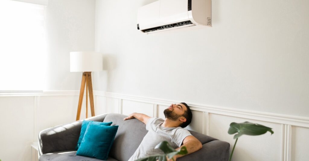 A person reclining on a gray sofa beneath a wall air conditioner, with a lamp, window, plant, and teal pillow.
