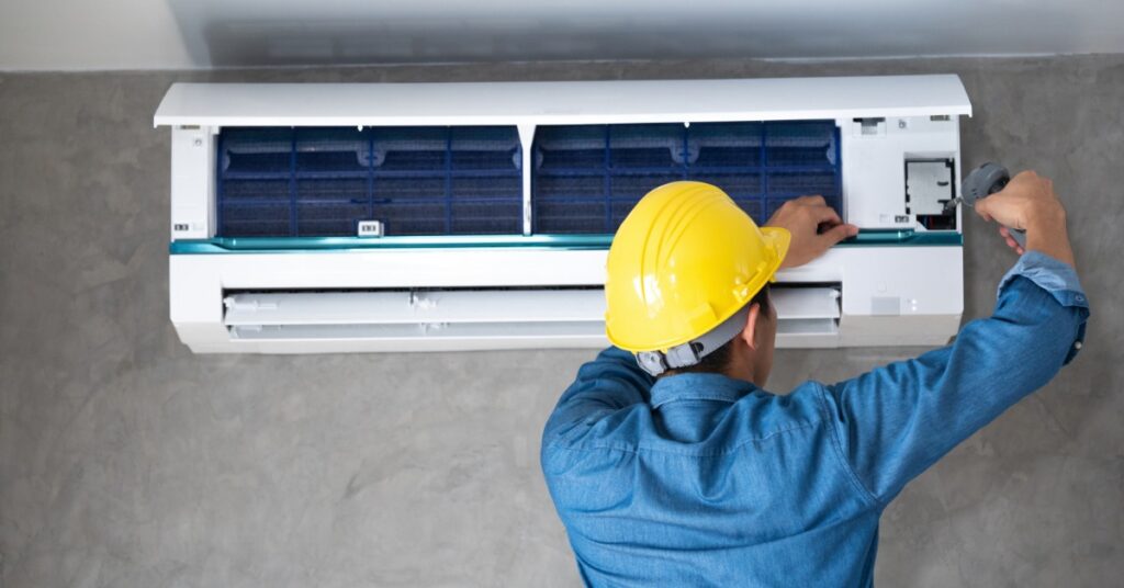 A person in a yellow hard hat and blue shirt using a screwdriver on an open wall air conditioner with a gray wall.