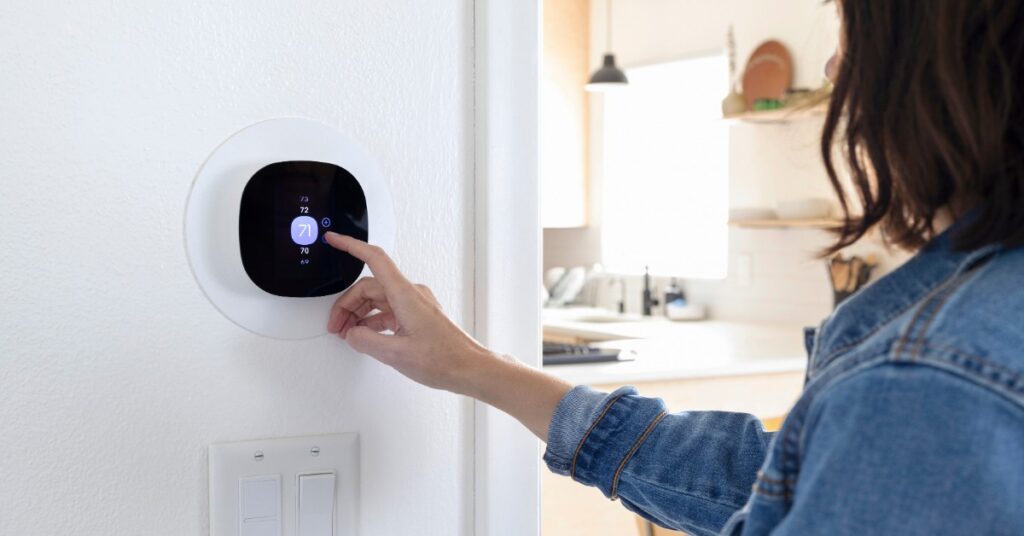 A woman in a denim shirt touching a round digital thermostat on a white wall beside two light switches.