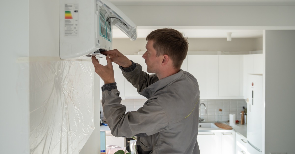 A man in a gray uniform adjusting and repairing a wall-mounted air conditioner mini-split system inside a home.