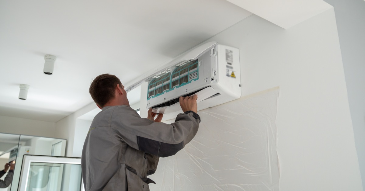 A man in a gray uniform working on an open wall-mounted air conditioner above plastic sheeting in a white room.